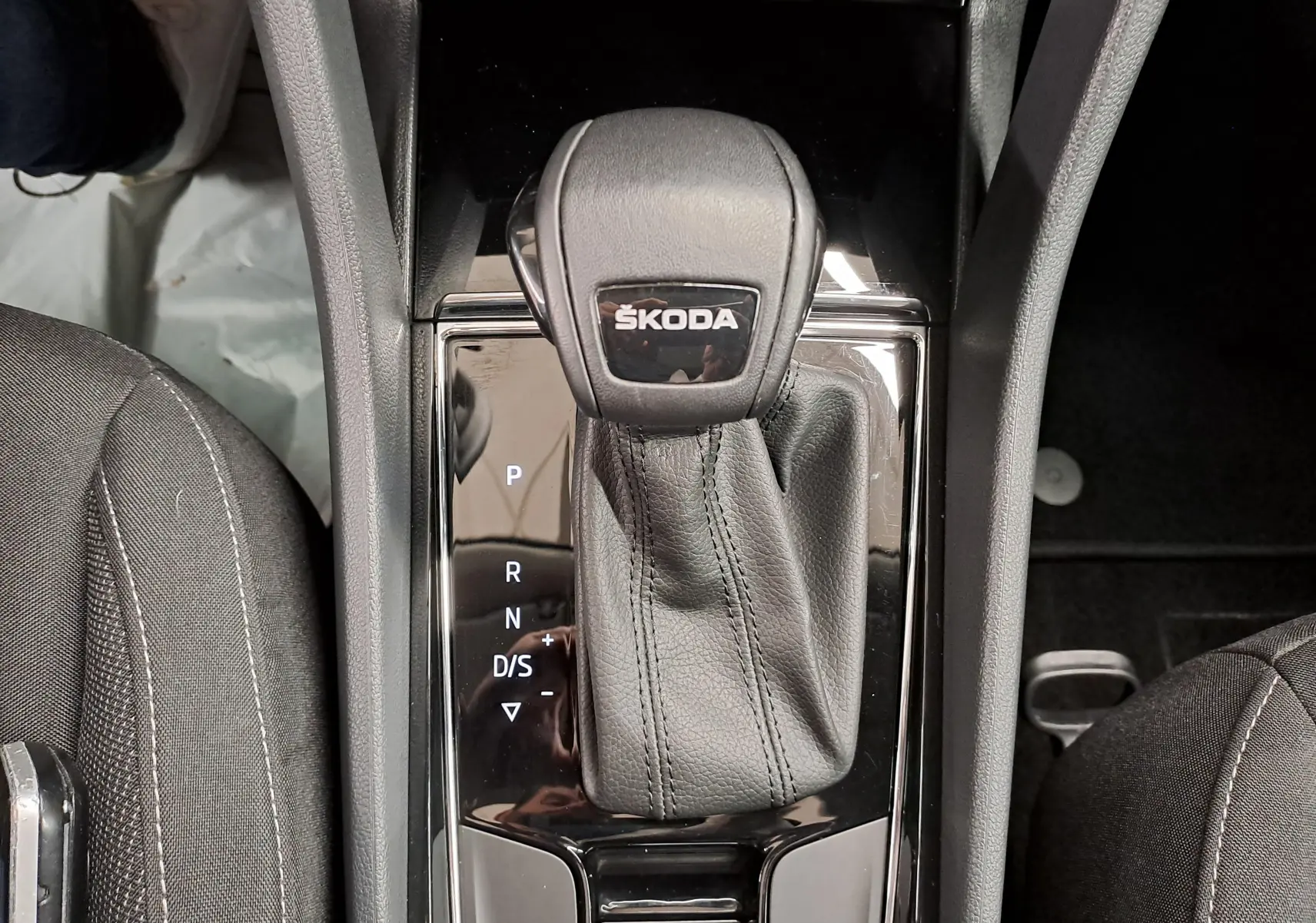 Gros plan sur le levier de vitesses automatique noir avec logo ŠKODA, entouré des sièges avant en tissu gris foncé.