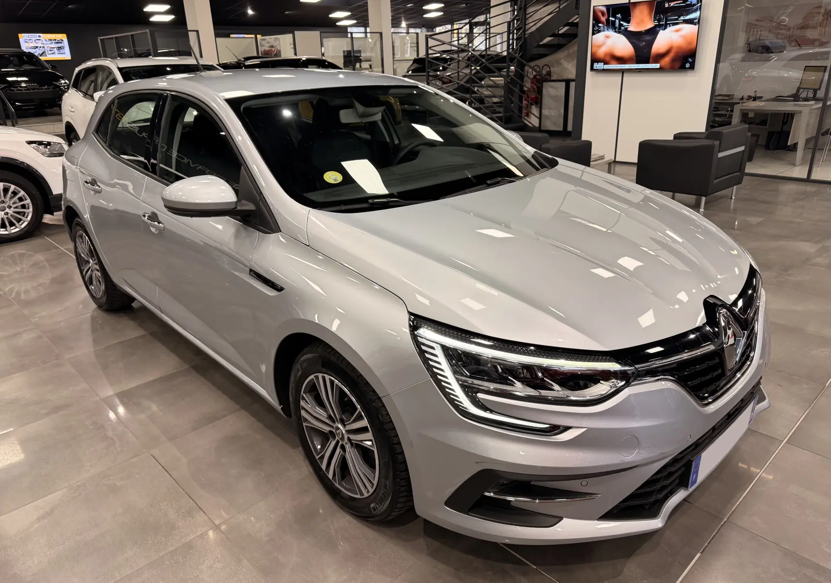 Renault Mégane IV gris Highland vue 3/4 avant droit en showroom, avec phares LED et jantes alliage.
