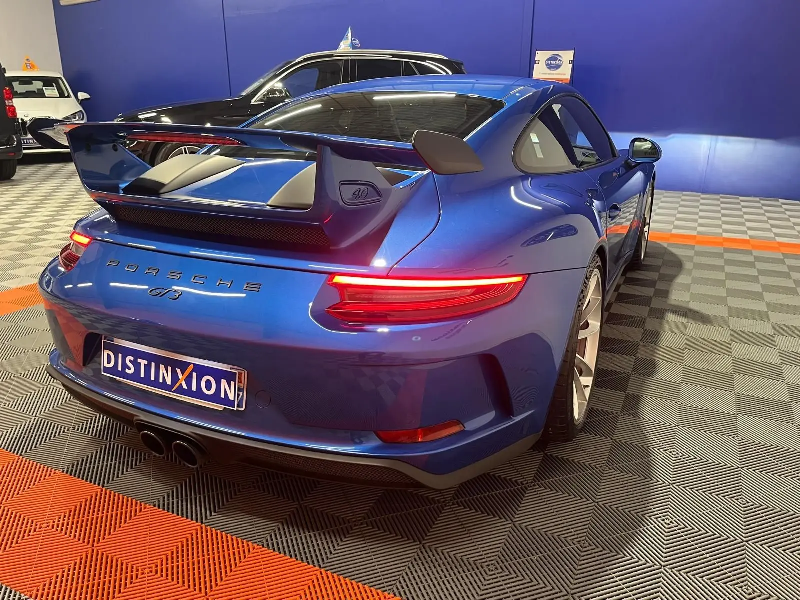 Vue 3/4 arrière droite d'une Porsche 911 GT3 bleu saphir avec grand aileron et feux arrière allumés.