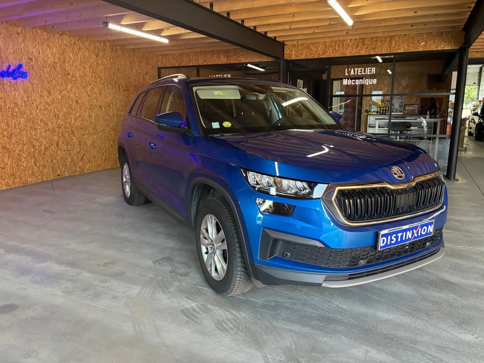Vue 3/4 avant d'un Skoda Kodiaq bleu 2023 avec calandre noire et jantes argentées, stationné en intérieur.