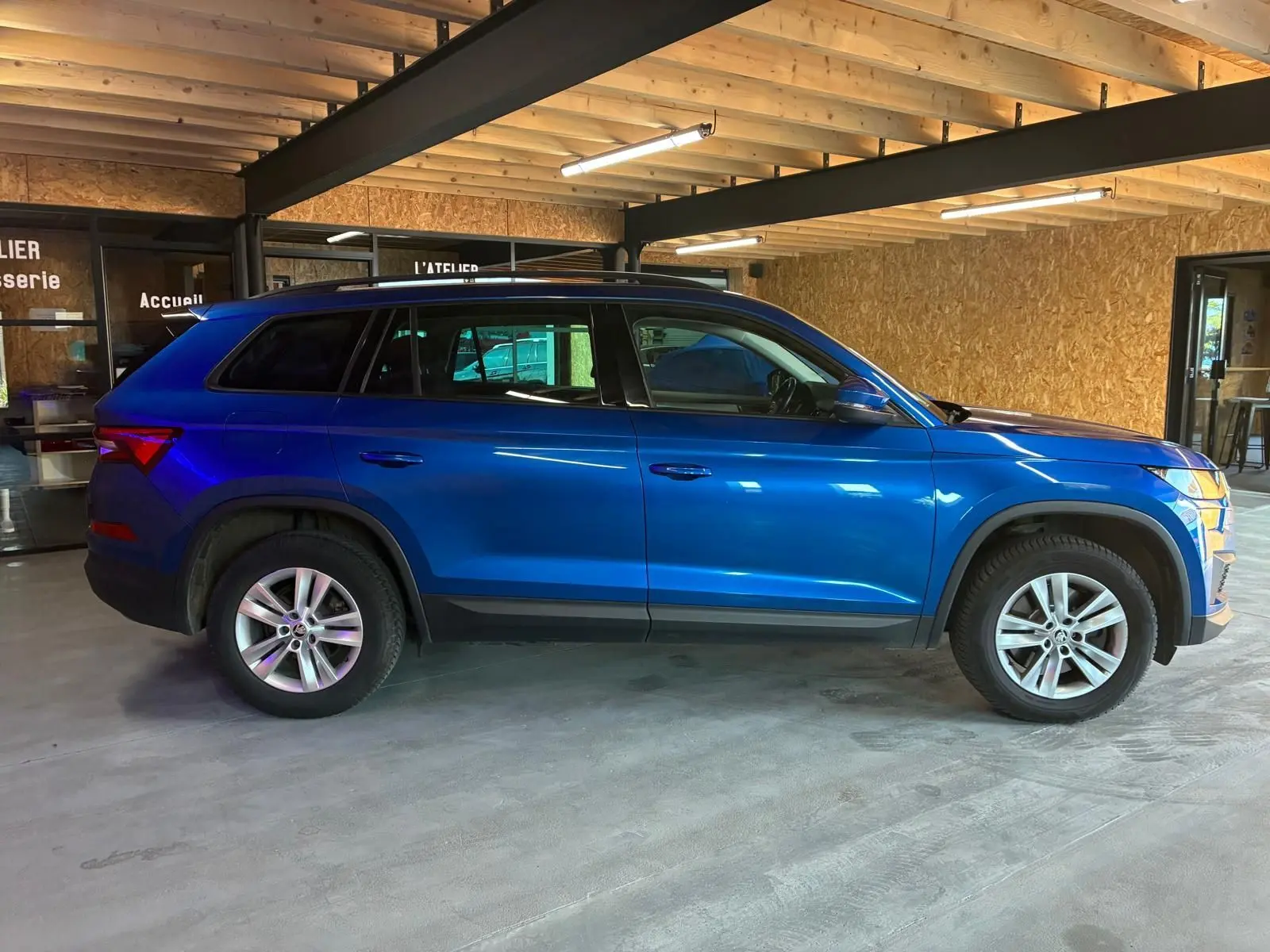 Vue de profil côté gauche d'un Skoda Kodiaq bleu 2023 garé en intérieur, soulignant ses lignes SUV et ses jantes alliage.