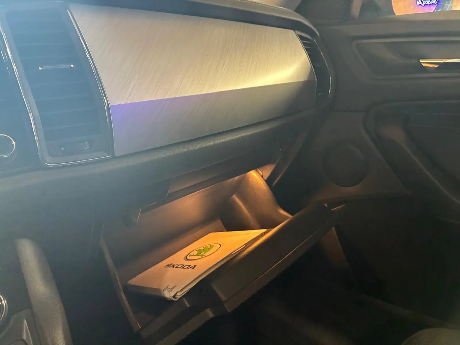Intérieur du Skoda Kodiaq 2023, vue côté passager sur la boîte à gants ouverte avec manuel Skoda visible, ambiance sombre.