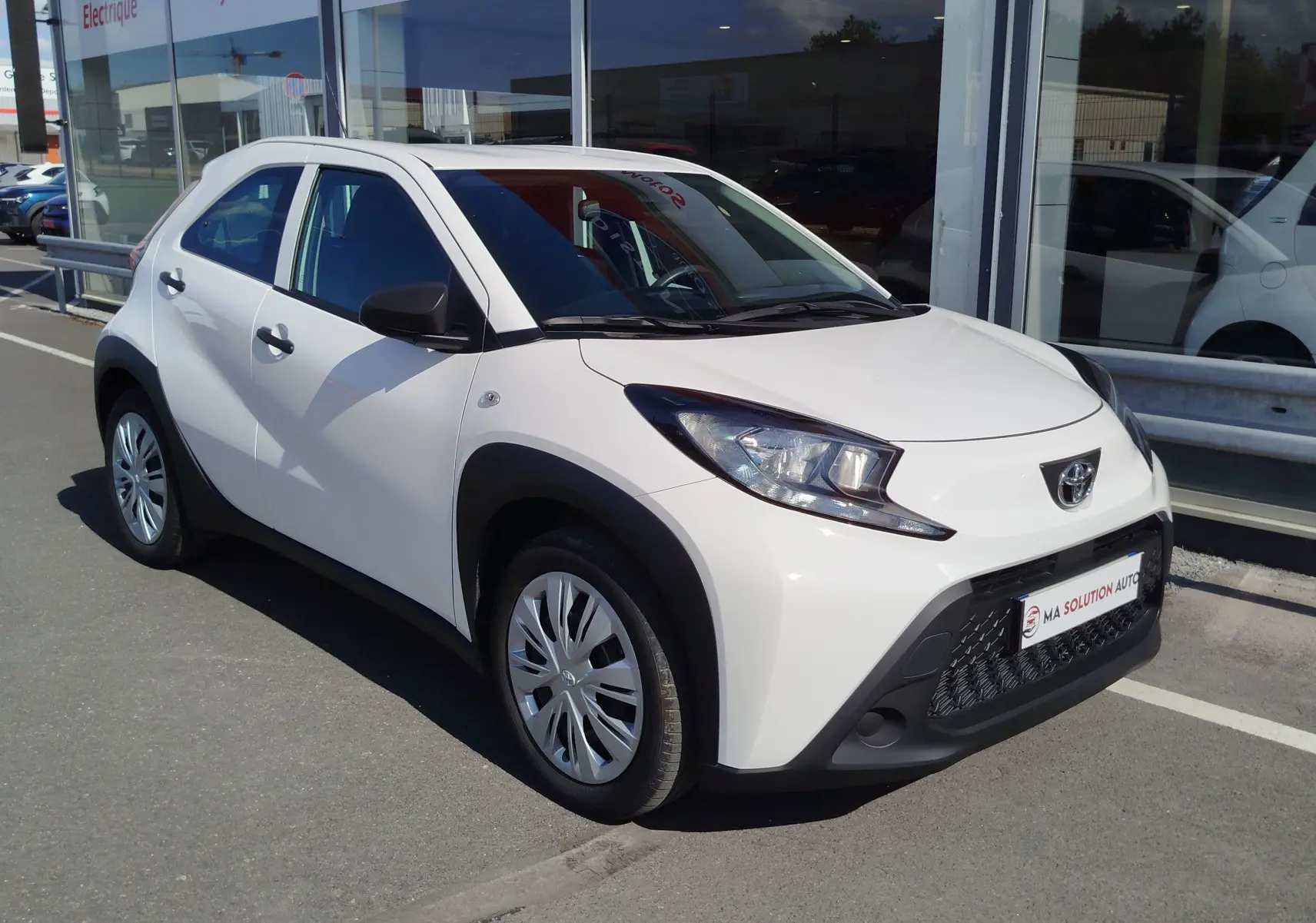 Toyota Aygo X blanc pur vue 3/4 avant droit, avec protections noires autour des passages de roues et calandre noire.