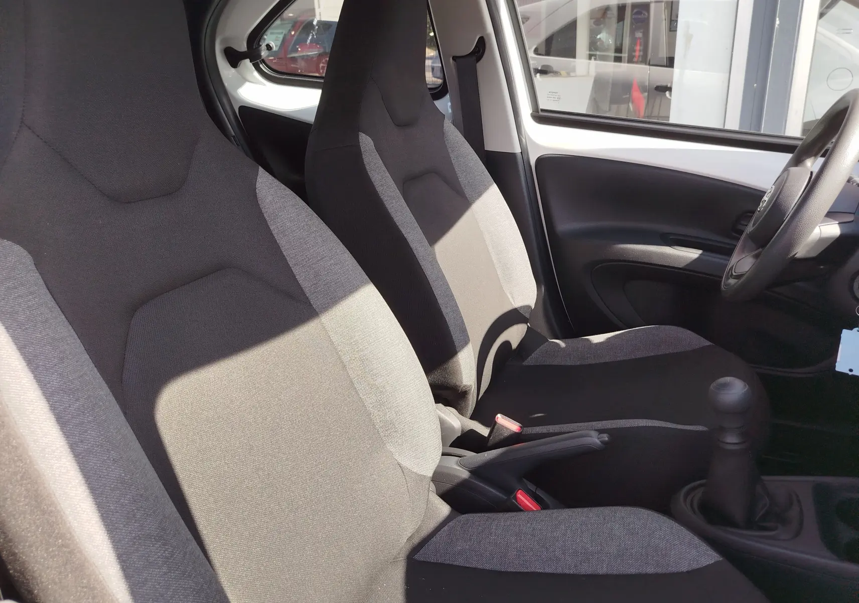 Vue intérieure côté conducteur de la Toyota Aygo X blanche, montrant les sièges tissu bicolore et la boîte manuelle.