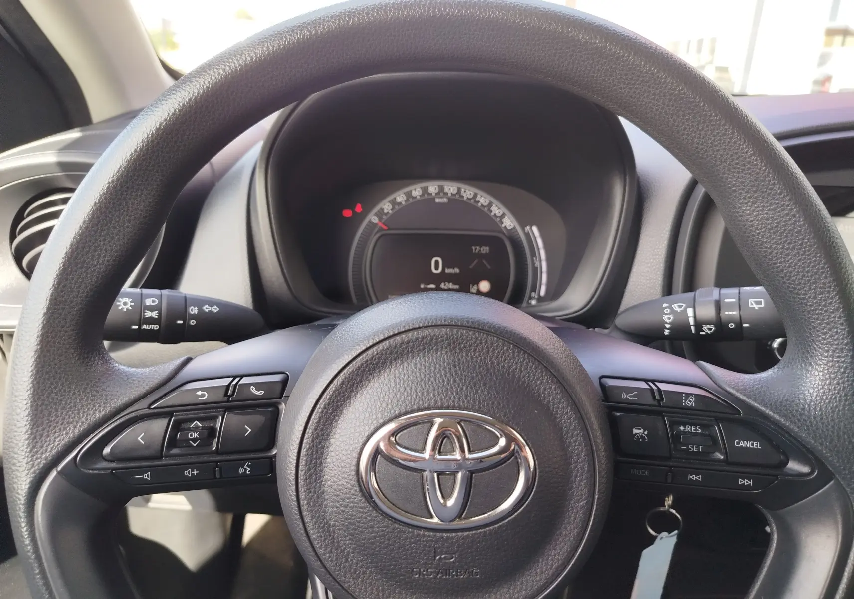 Vue rapprochée du volant noir de la Toyota Aygo X 2023, avec commandes multifonctions et tableau de bord digital en arrière-plan.