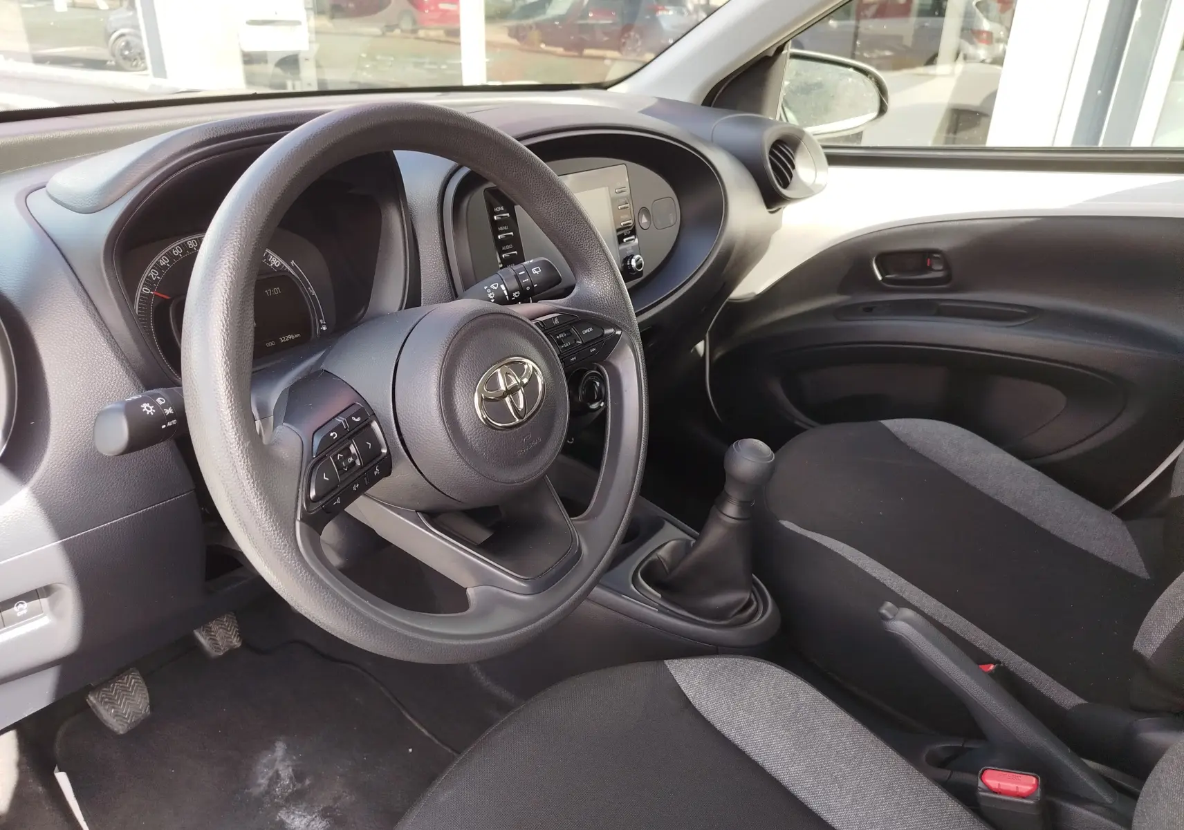 Vue intérieure côté conducteur de la Toyota Aygo X 2023, volant multifonction, sièges tissu noir et gris, levier de vitesse manuel.