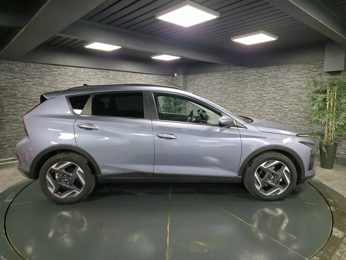 Vue de profil côté gauche d'une Hyundai Bayon gris métallisé 2025 avec jantes alliage et vitres surteintées dans un showroom.