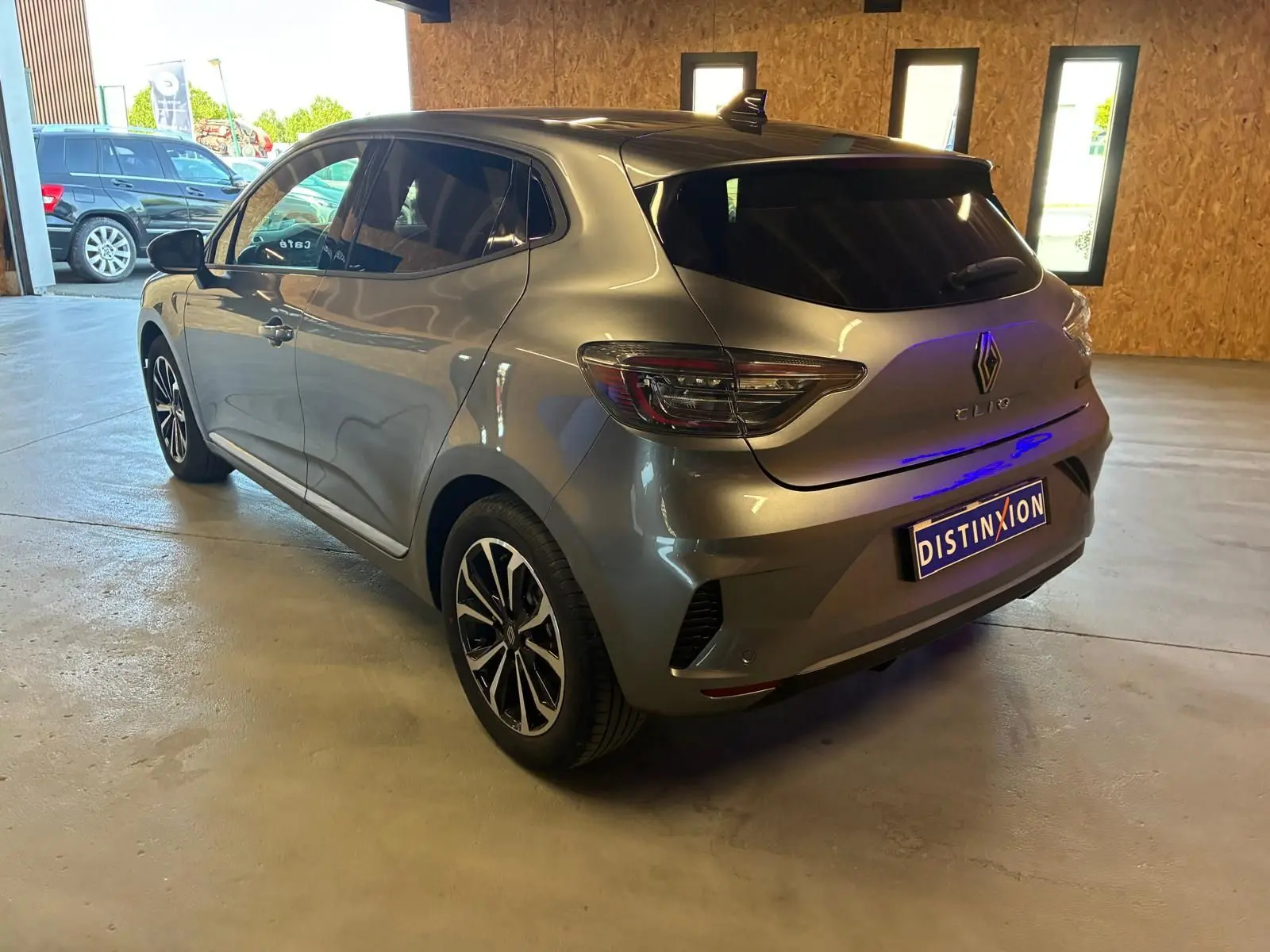 Vue 3/4 arrière droite d'une Renault Clio gris schiste, avec feux LED et jantes alliage noires, en intérieur lumineux.