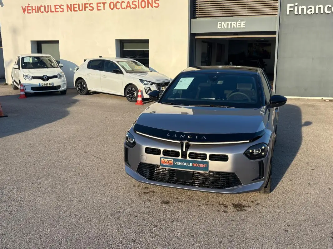 Lancia Ypsilon gris vue de face, stationnée devant un bâtiment avec panneau "Véhicules neufs et occasions".