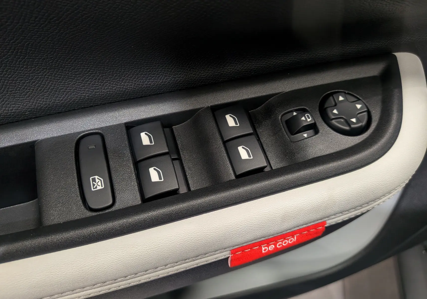 Gros plan sur les commandes de lève-vitres et rétroviseurs électriques du côté gauche, garniture cuir clair avec étiquette rouge "be cool" visible.