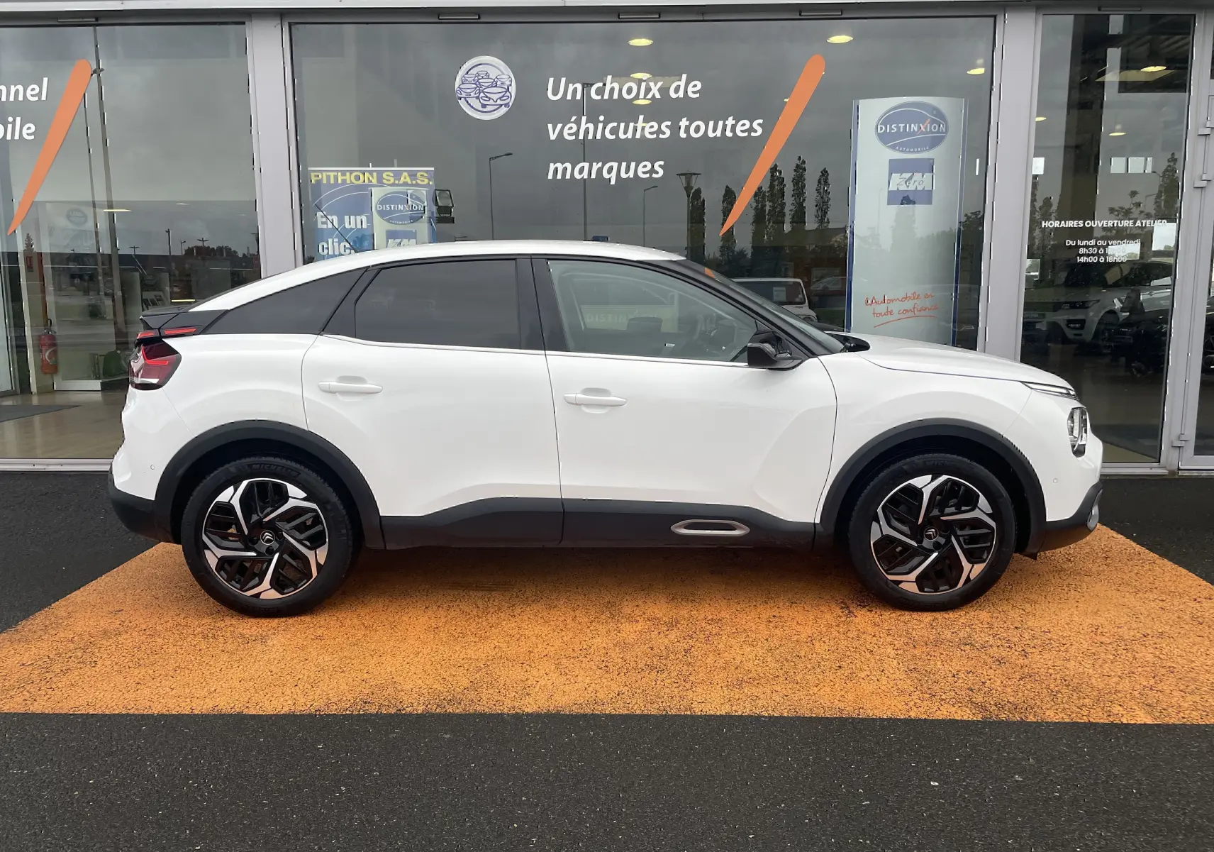 Profil droit d'une Citroën C4 blanche 2022 avec toit noir et jantes alliage distinctives, stationnée devant une vitrine.