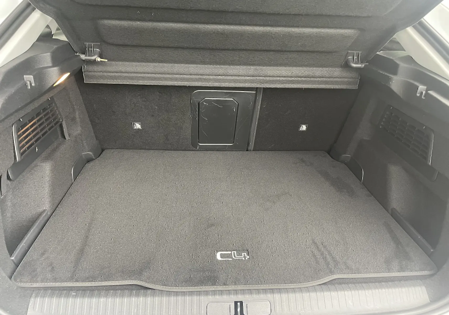 Coffre ouvert vu de l'arrière d'une Citroën C4 blanche, tapis noir avec logo C4 et éclairage intérieur.