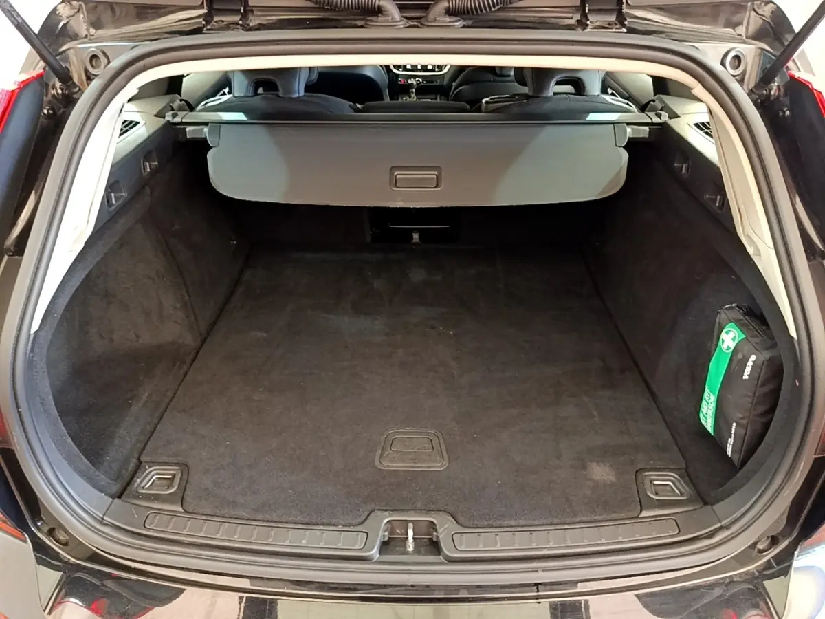 Coffre ouvert vue arrière d’un Volvo V60 noir, avec cache-bagages et trousse de secours visible à droite.