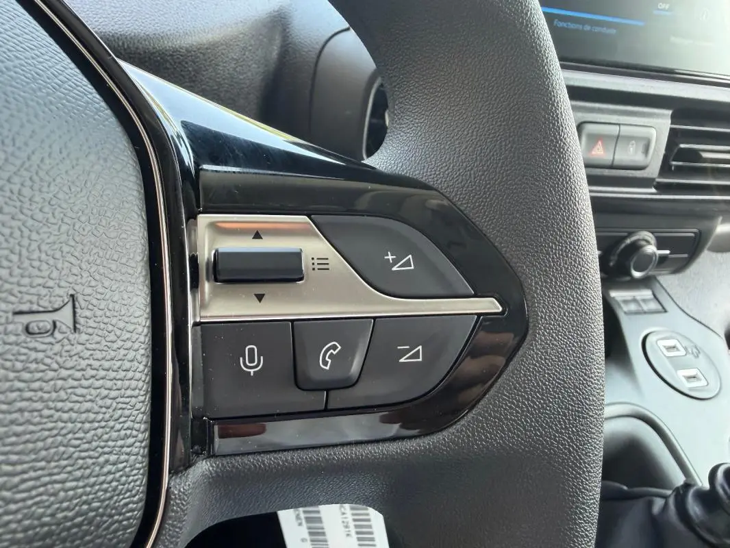 Gros plan sur les commandes du volant cuir noir du Peugeot Partner blanc, avec boutons multifonctions et tableau de bord en arrière-plan.