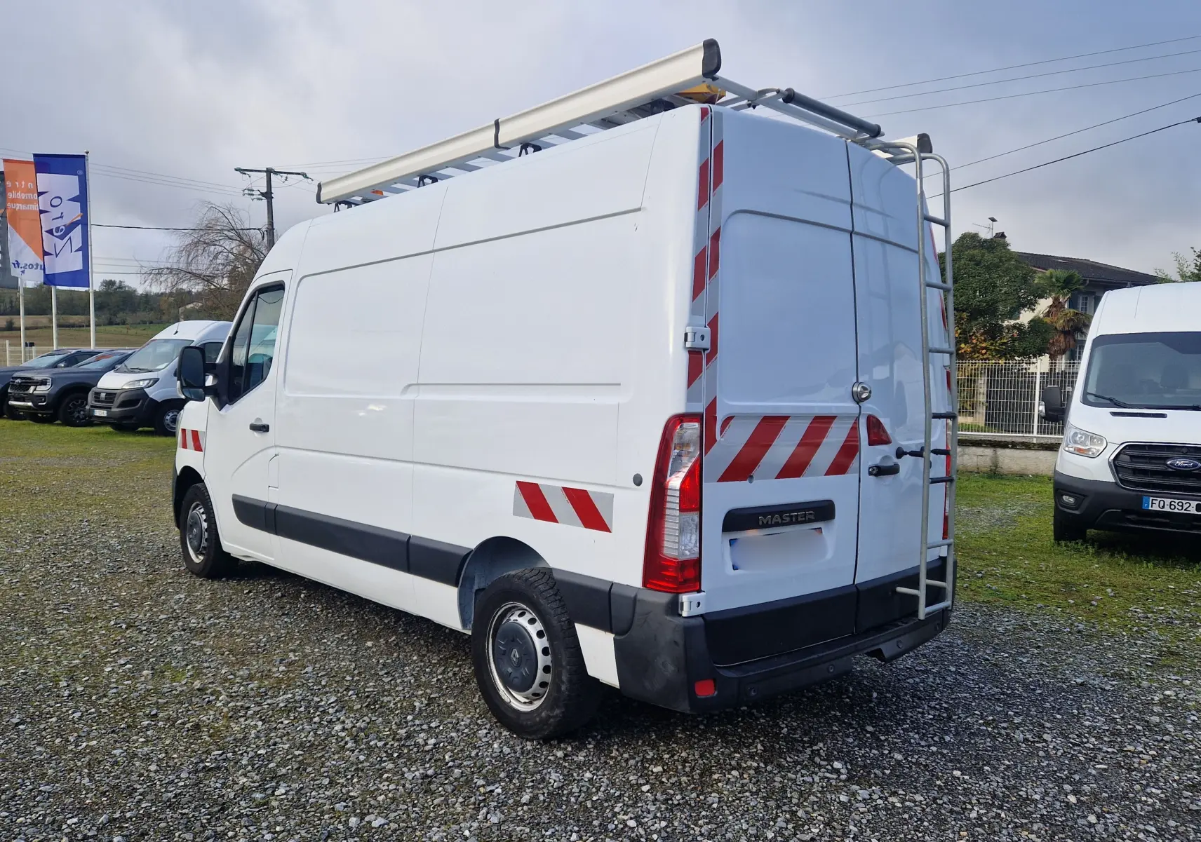 Renault Master blanc en vue 3/4 arrière droit, avec galerie de toit, échelle arrière et bandes réfléchissantes rouges.