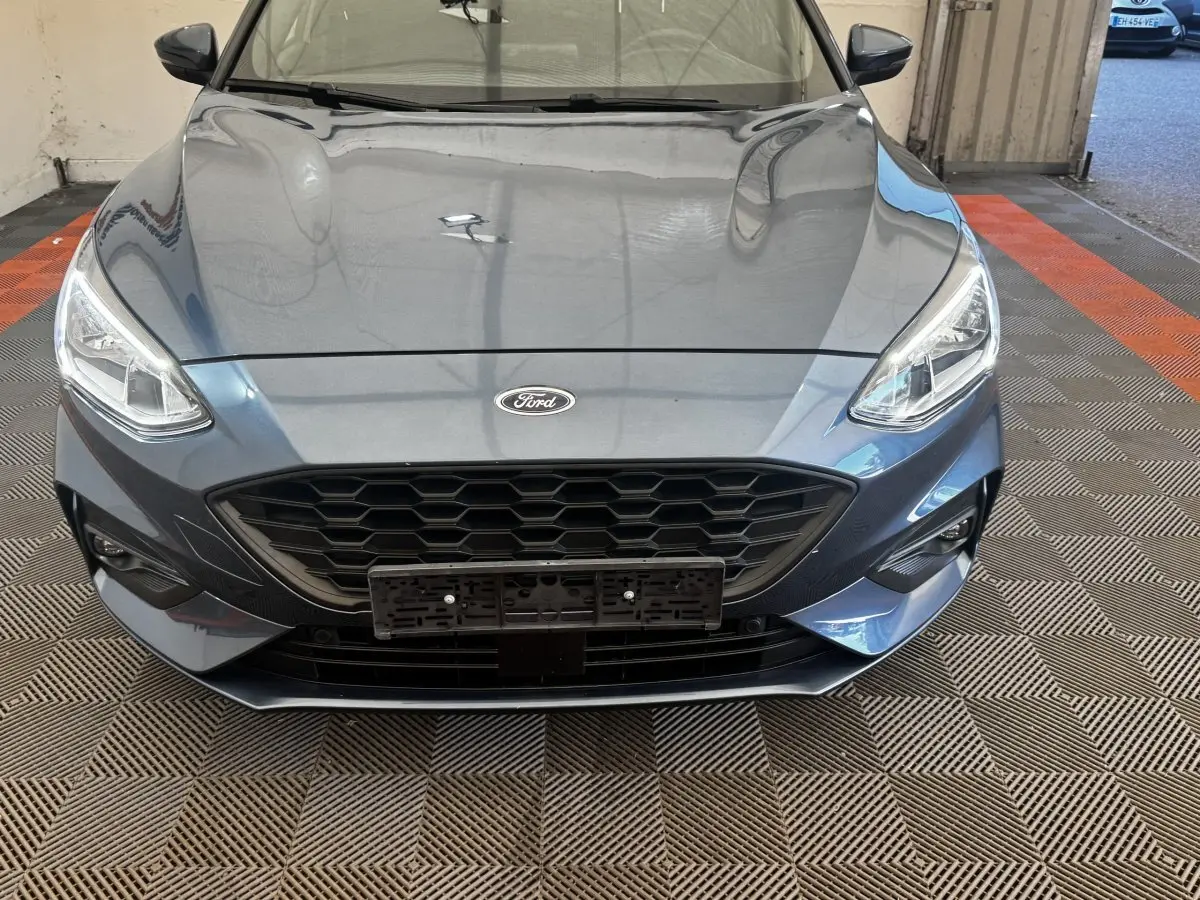 Vue avant rapprochée d'une Ford Focus 5P St-Line bleu métallisé, mettant en valeur sa calandre hexagonale et ses phares LED.