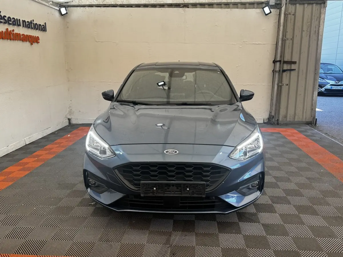 Vue frontale d'une Ford Focus 5 portes bleu métallisé avec calandre noire et phares allumés.
