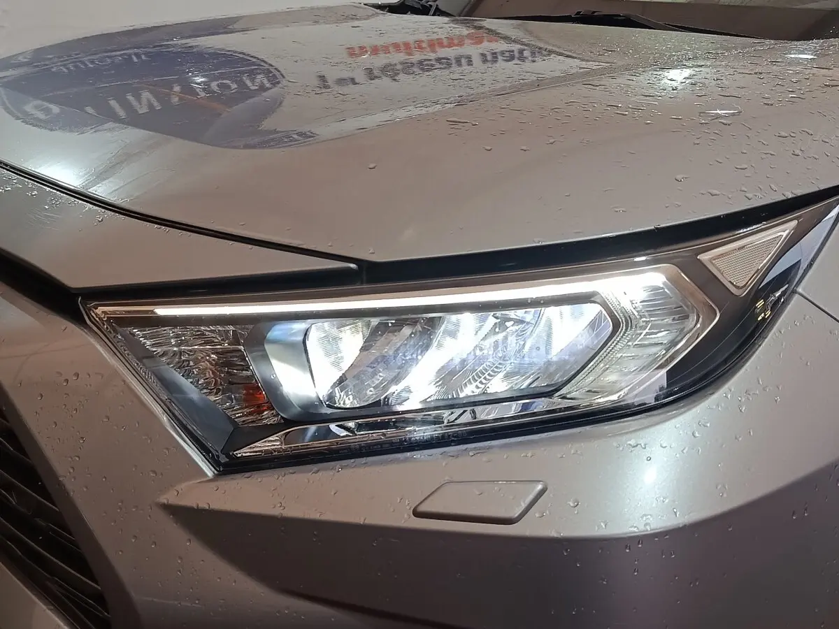 Gros plan sur le phare avant droit allumé d’un Toyota RAV4 hybride gris clair avec gouttes d’eau sur la carrosserie.