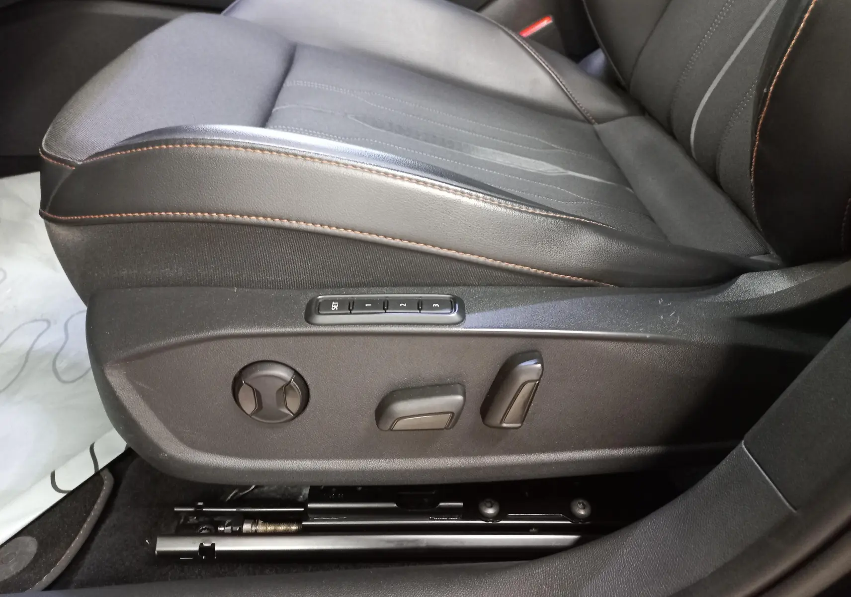 Gros plan sur le siège conducteur noir avec surpiqûres orange et commandes électriques de réglage latérales.