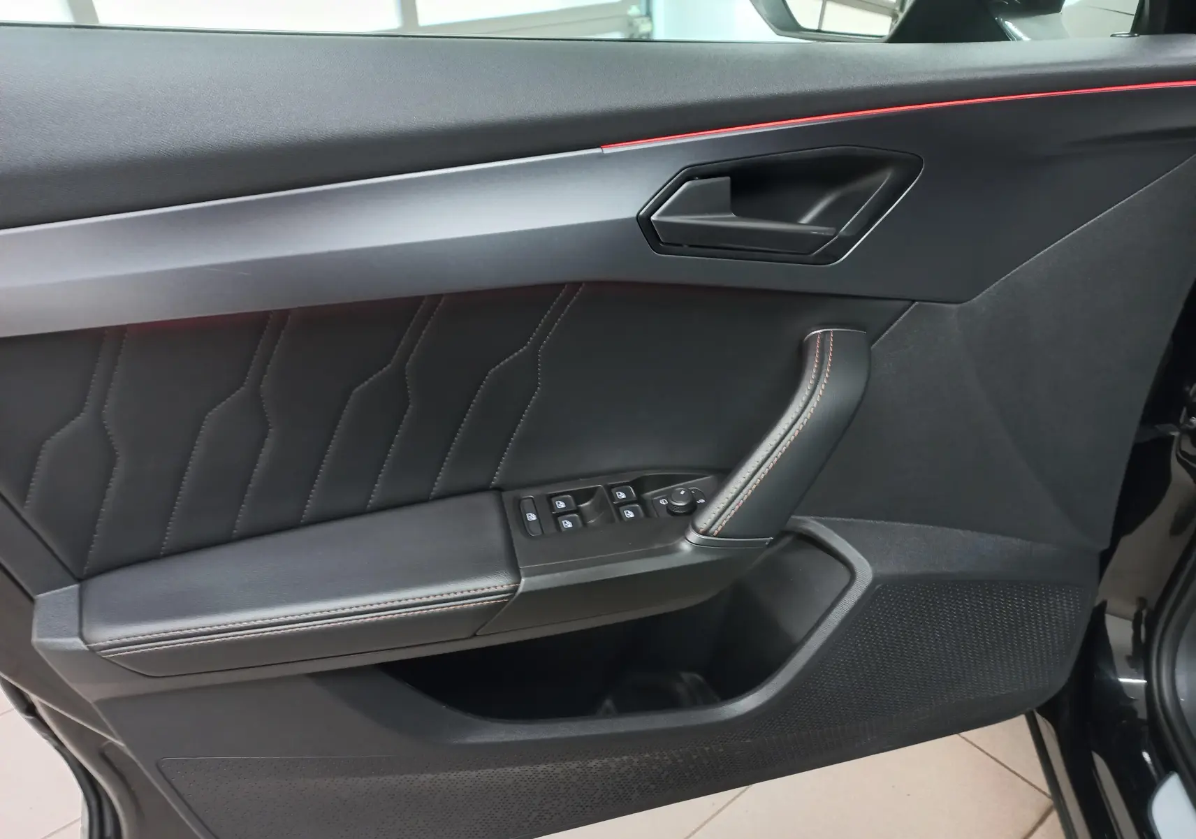 Vue intérieure côté gauche de la porte d'une CUPRA Formentor noire avec surpiqûres orange et éclairage d'ambiance rouge.