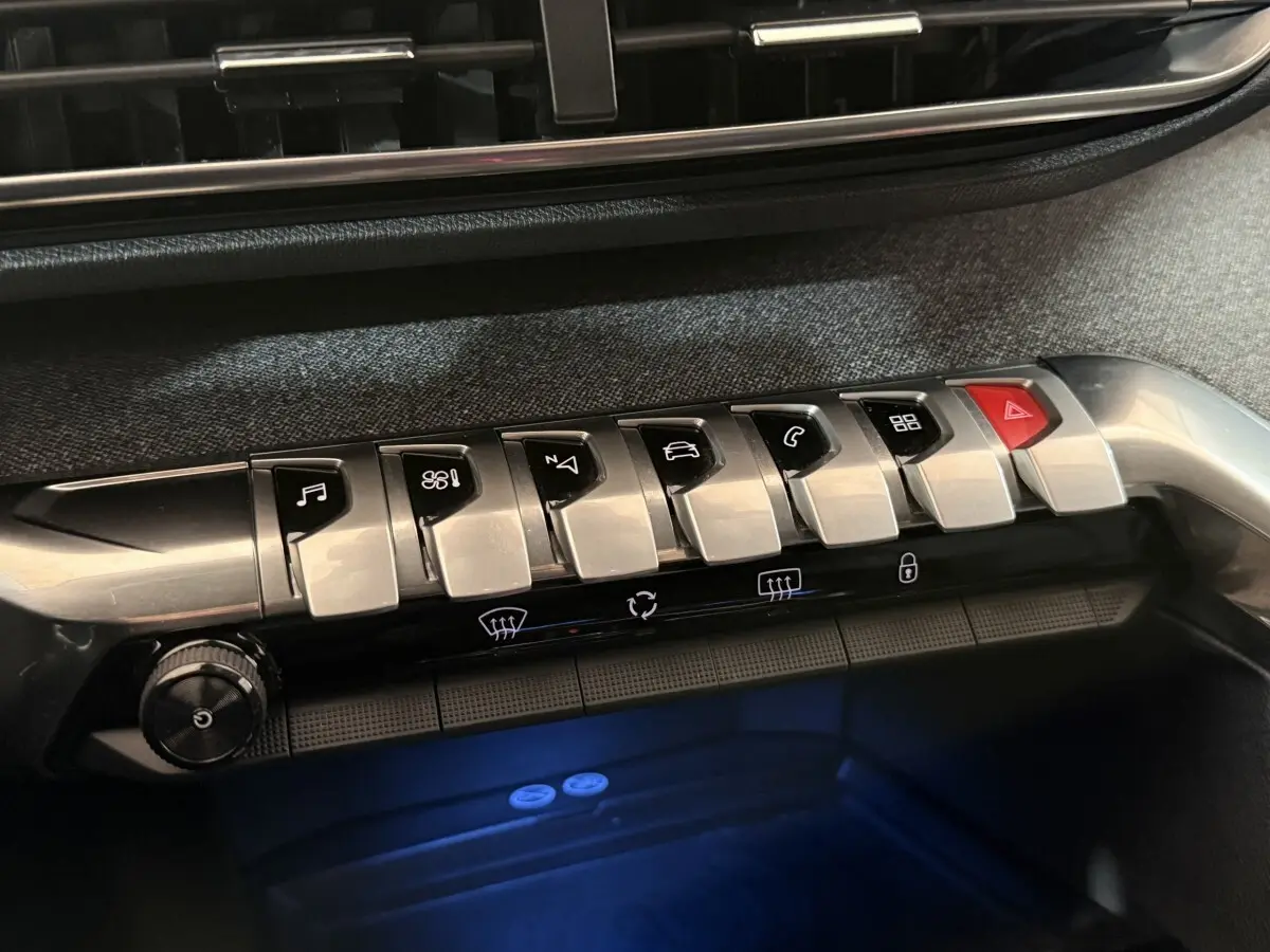 Gros plan sur la rangée de boutons argentés et noirs du tableau de bord du Peugeot 5008, éclairage bleu en dessous.