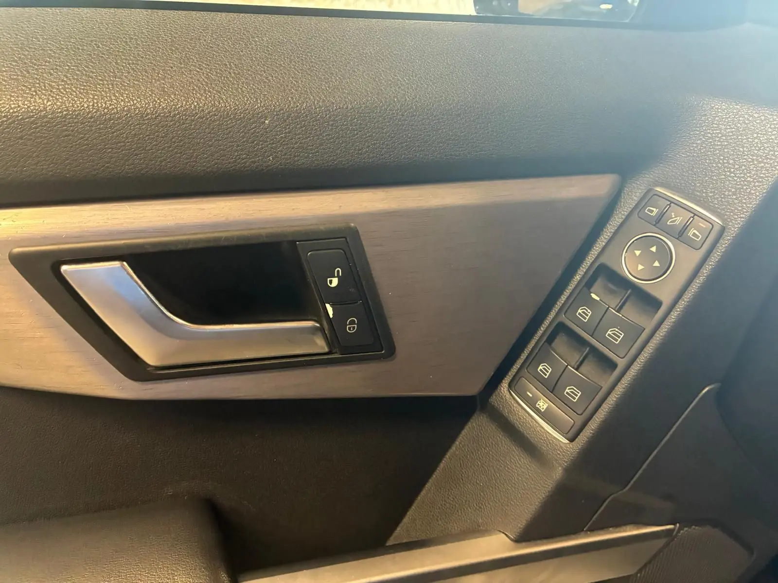 Gros plan sur la poignée de porte côté gauche et les commandes de vitres électriques d’un Mercedes GLK noir, intérieur bois et cuir.