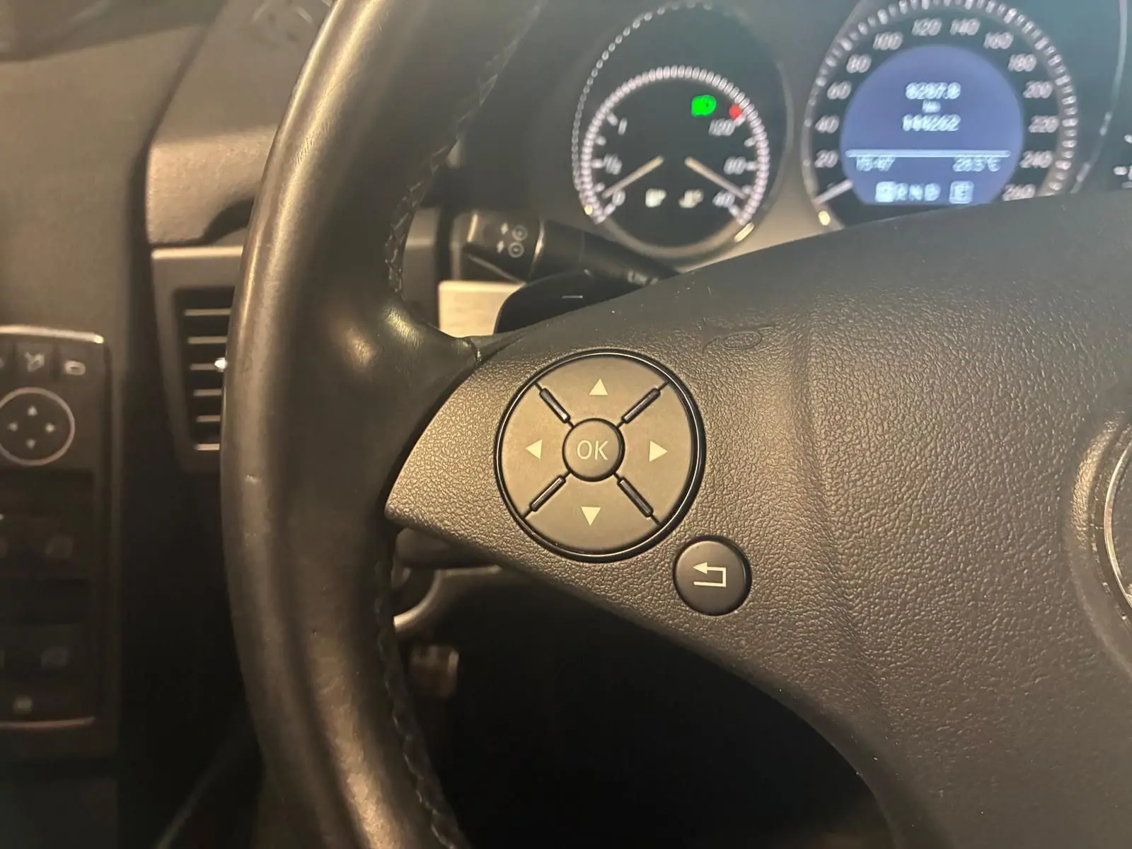 Gros plan sur le volant en cuir noir d’un Mercedes GLK 220 CDI, avec commandes intégrées et tableau de bord éclairé en arrière-plan.
