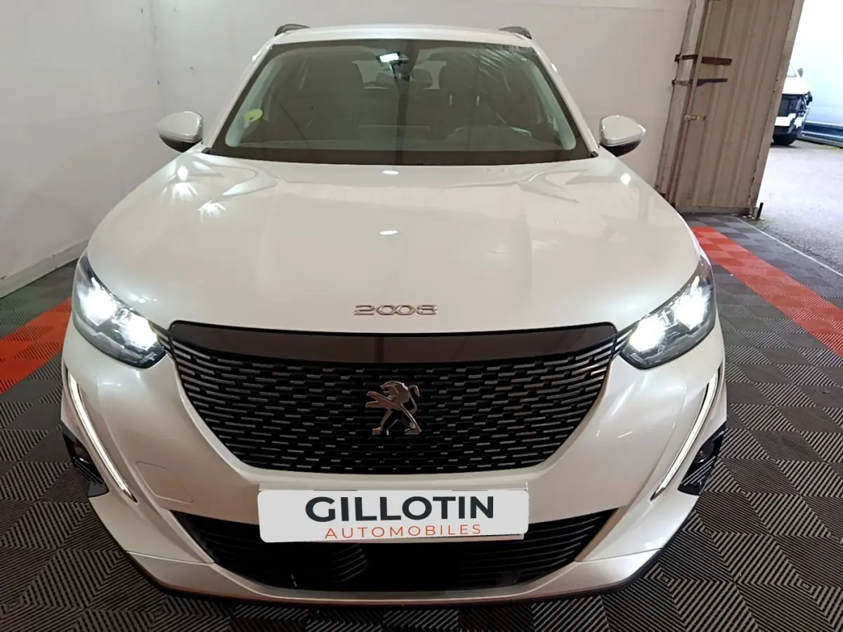 Vue avant d'un Peugeot 2008 blanc avec calandre noire et phares LED allumés dans un garage.