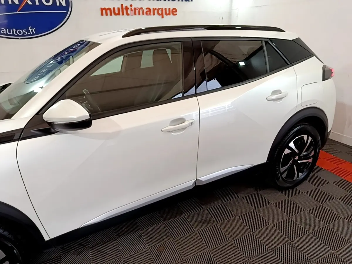 Vue latérale droite d’un Peugeot 2008 blanc avec toit noir et jantes alliage distinctives en intérieur showroom.