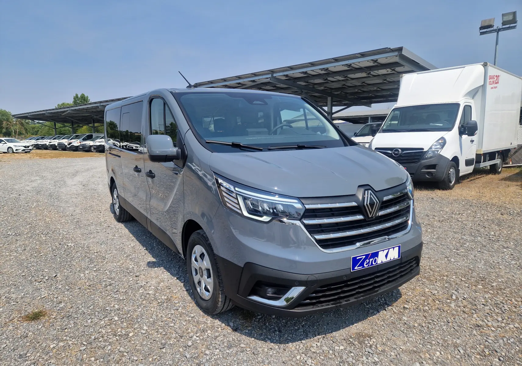 Vue 3/4 avant droit d’un Renault Trafic Combi gris urbain stationné sur un sol gravillonné avec un abri solaire en arrière-plan.