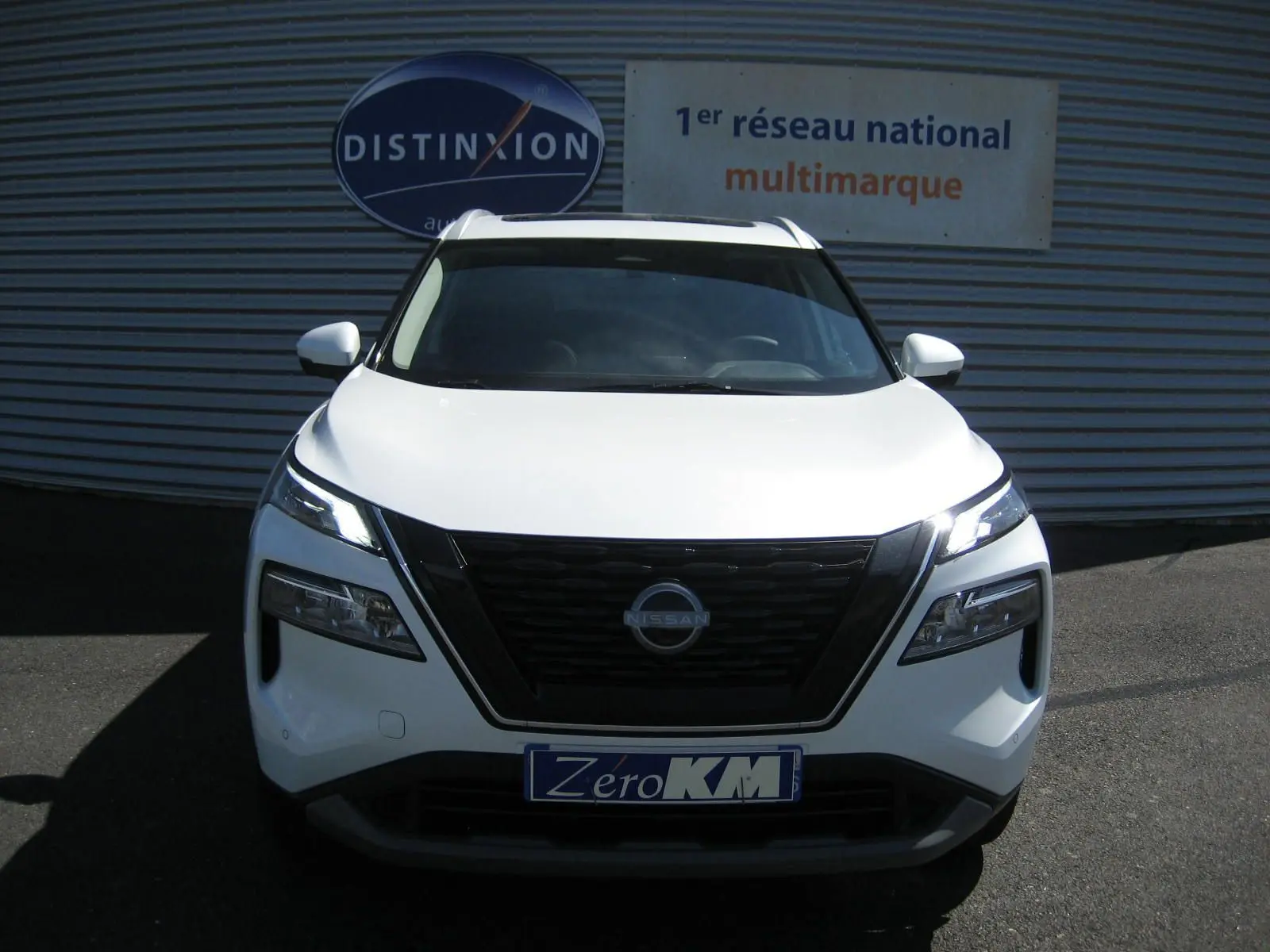 Vue de face du Nissan X-TRAIL blanc nacré avec calandre noire et phares LED allumés sur fond de façade commerciale.