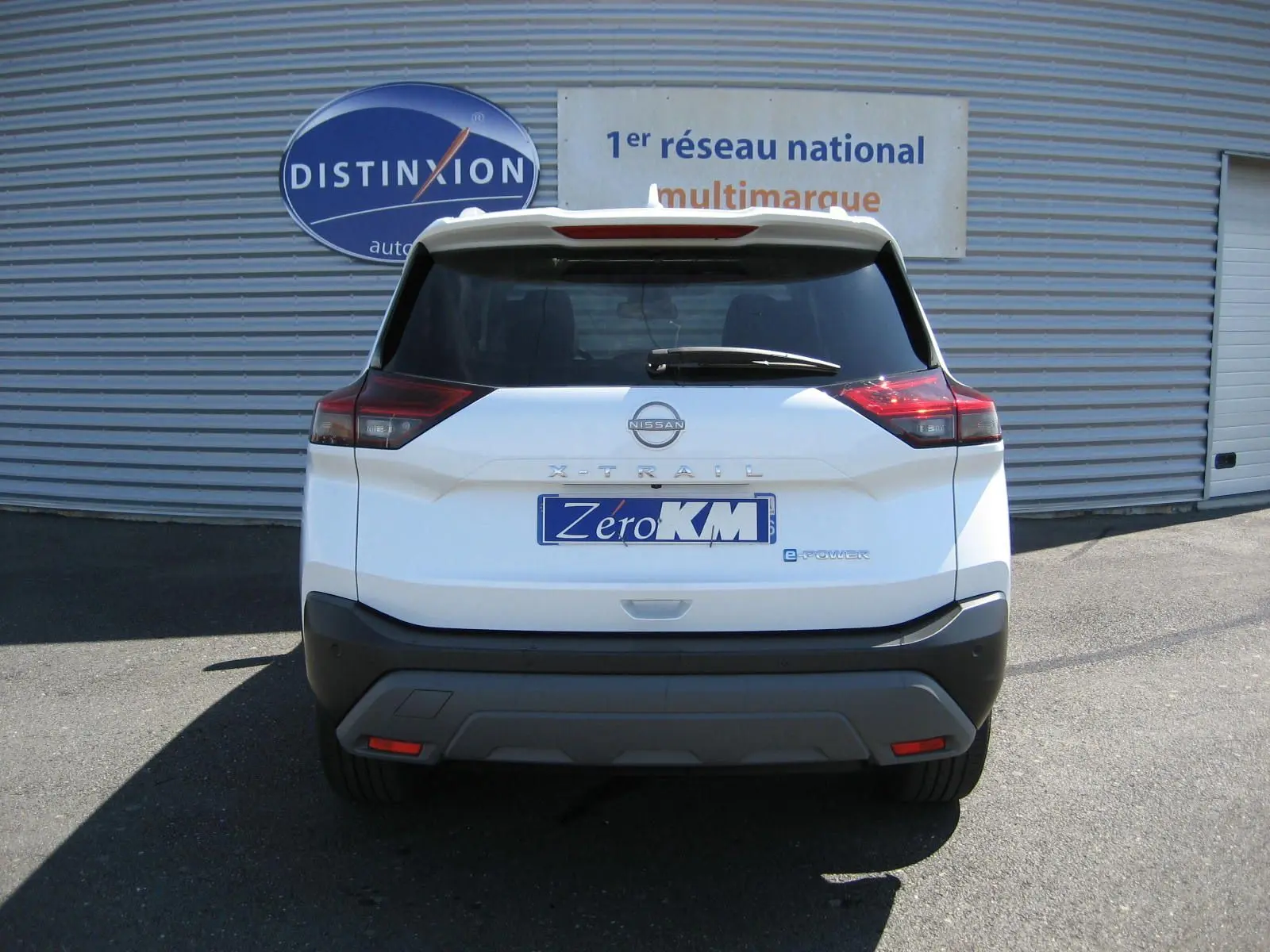 Vue arrière d'un Nissan X-Trail blanc nacré avec logo e-Power et plaque "Zéro KM" devant un mur métallique.
