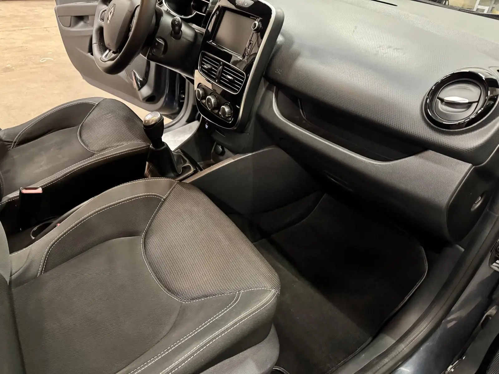 Intérieur avant droit d'une Renault Clio IV 2019, sièges tissu noir et tableau de bord gris foncé avec boîte manuelle.