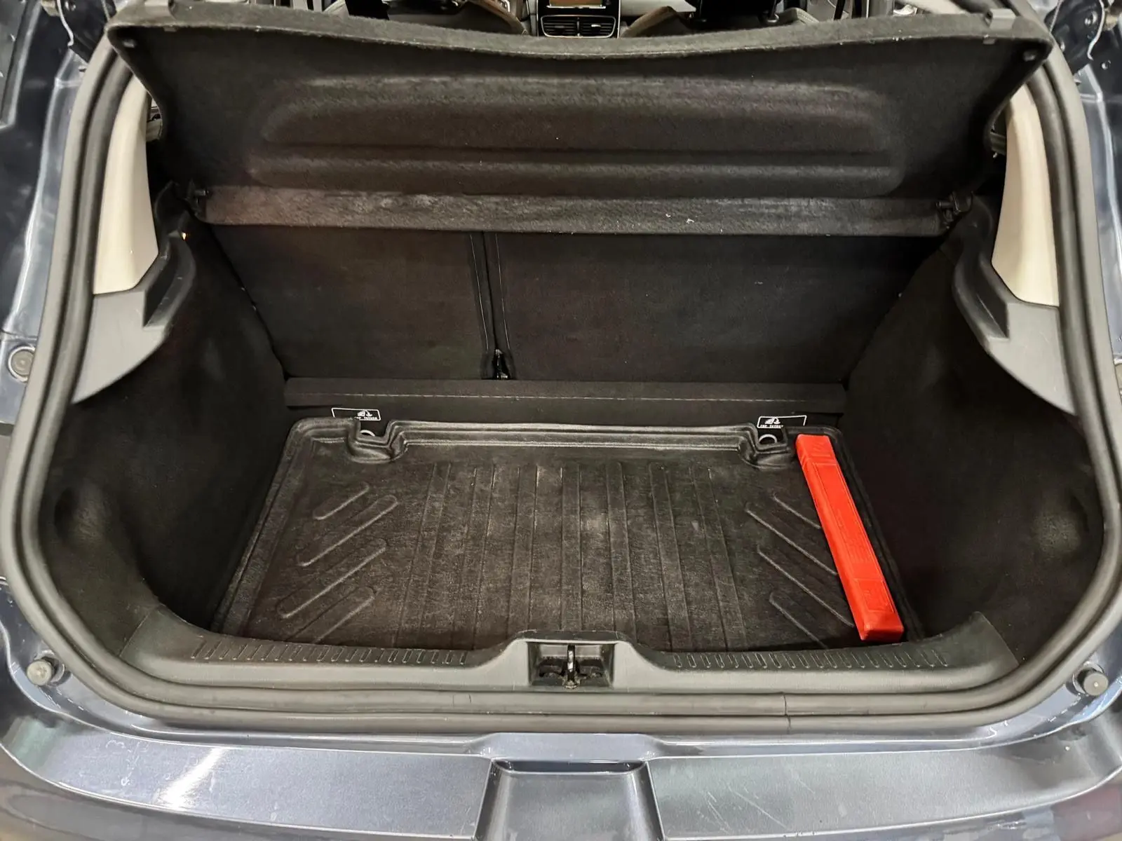 Coffre ouvert d’une Renault Clio IV gris foncé, montrant un tapis de coffre noir avec un triangle de sécurité rouge.
