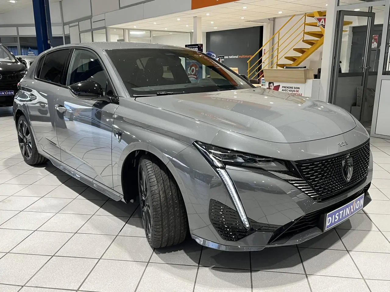 Peugeot 308 Gris Selenium vue 3/4 avant droit, calandre noire vernie et feux LED griffés en showroom.