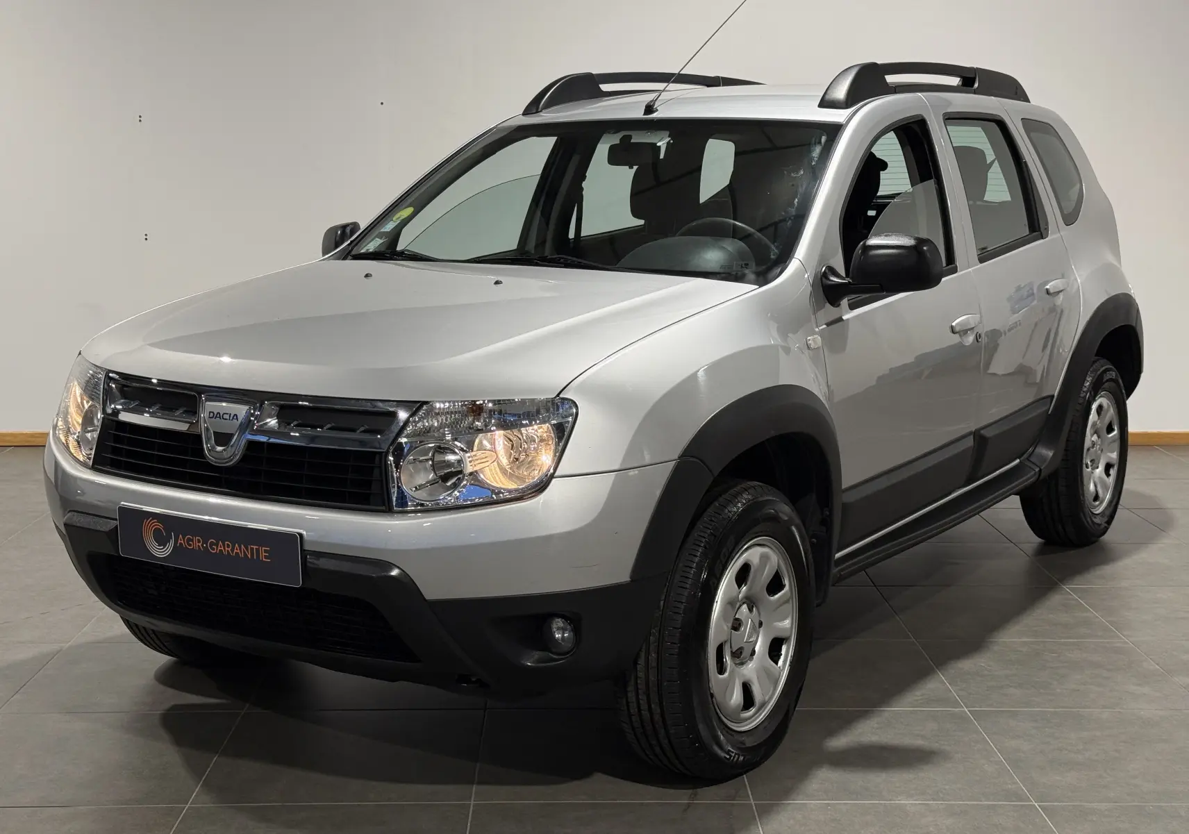 Dacia Duster gris en 3/4 avant droit avec attelage, phares allumés et protections noires autour des roues.