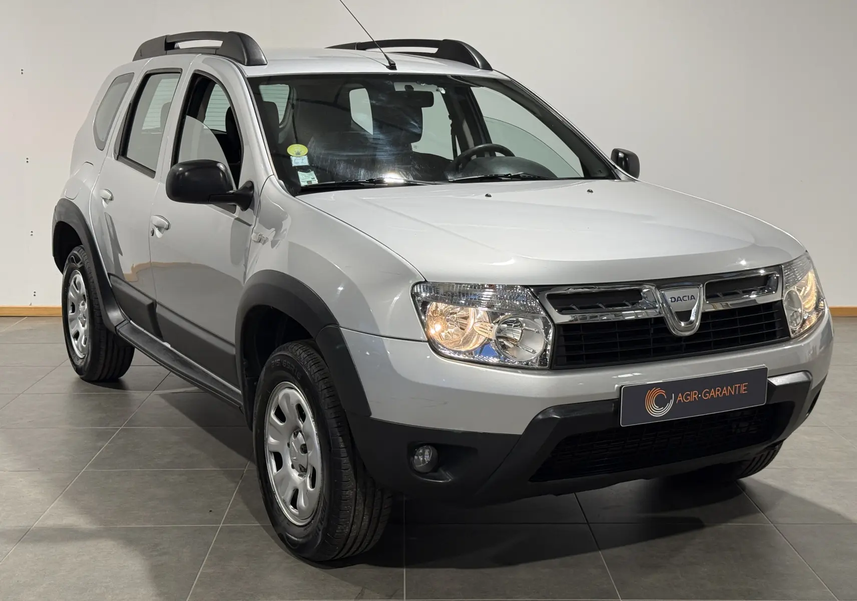 Vue 3/4 avant droit d'un Dacia Duster gris 2011 avec attelage et protections noires sur les passages de roues.