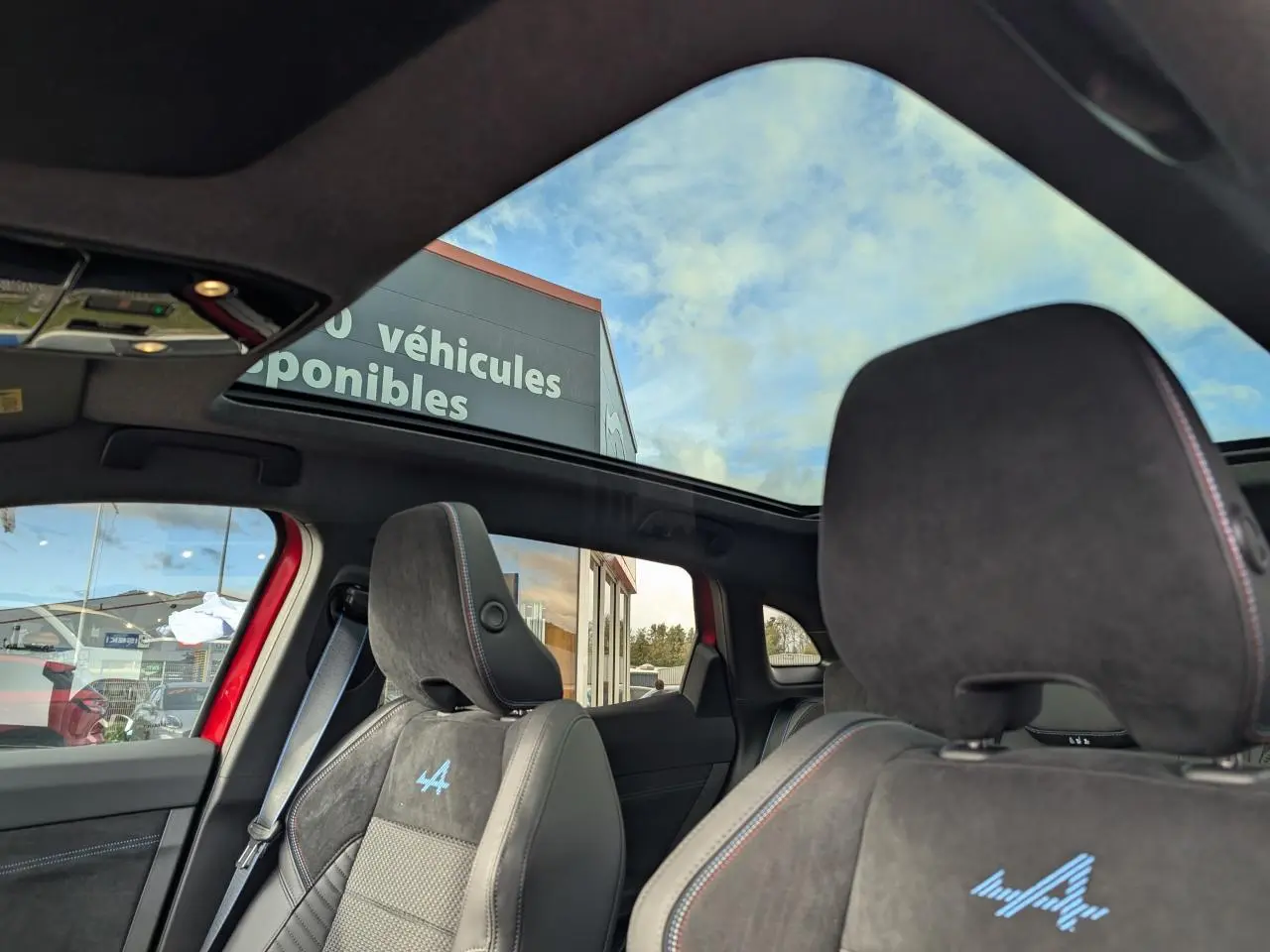 Vue intérieure du toit panoramique et des sièges avant en Alcantara noir avec logo Alpine bleu sur un Renault Austral rouge métallisé.