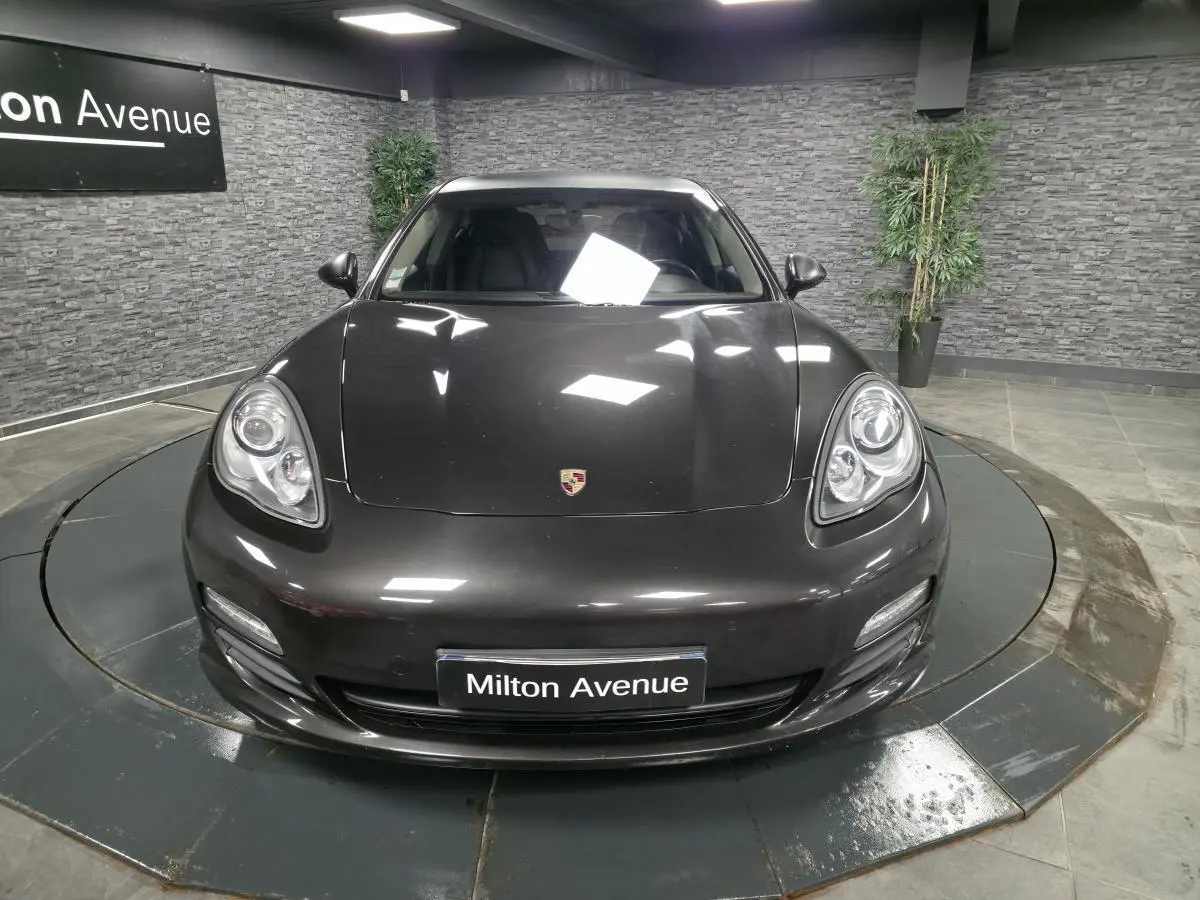 Vue avant d'une Porsche Panamera marron métallisé avec phares ovales et plaque Milton Avenue en intérieur showroom.
