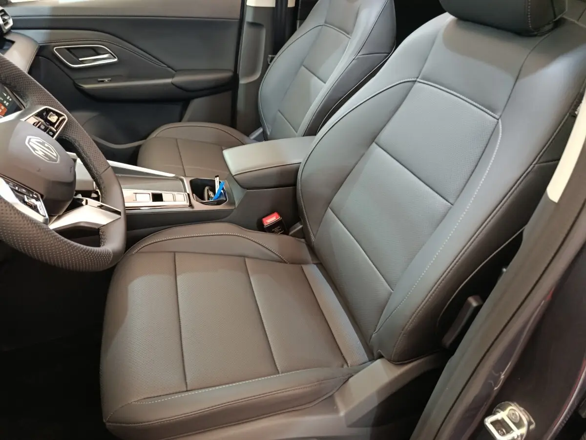 Vue intérieure côté conducteur du MG Motor ZS 1.5L Hybrid+ Luxury 2026, sièges cuir gris foncé avec accoudoir central.