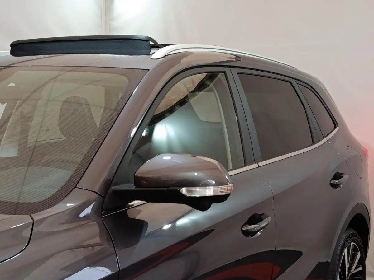 Vue de côté droit avant d’un MG ZS gris foncé avec rétroviseur et barres de toit visibles dans un intérieur neutre.