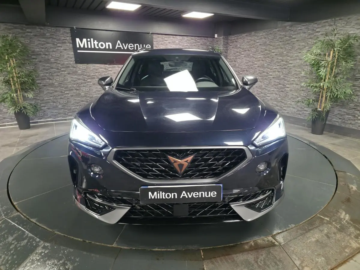 Vue frontale d'un CUPRA Formentor bleu avec calandre noire et logo cuivre, phares LED allumés dans un showroom.