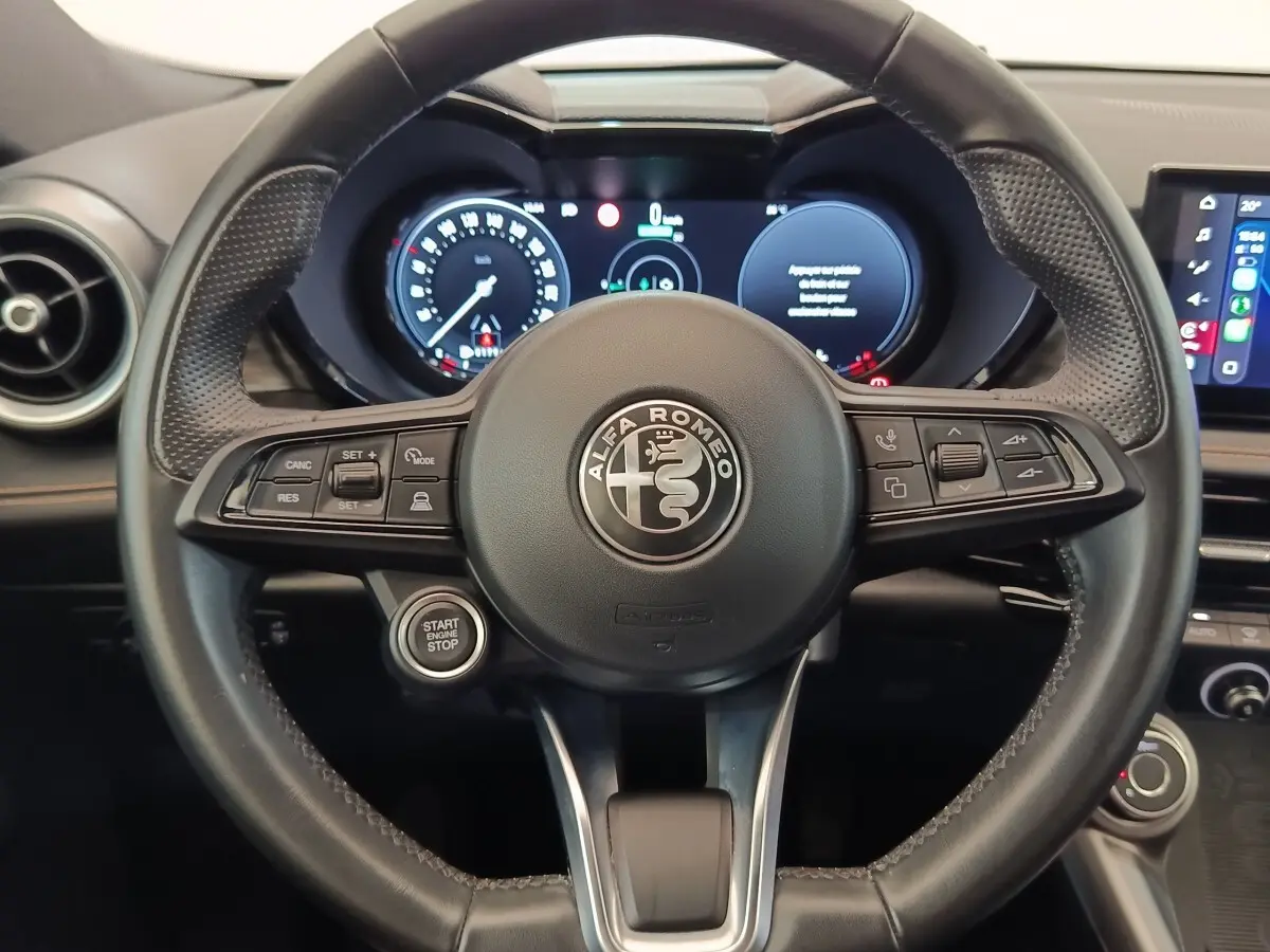 Vue rapprochée du volant en cuir noir de l'Alfa Romeo Tonale 1.5T Hybrid 160 VGT avec tableau de bord digital allumé.