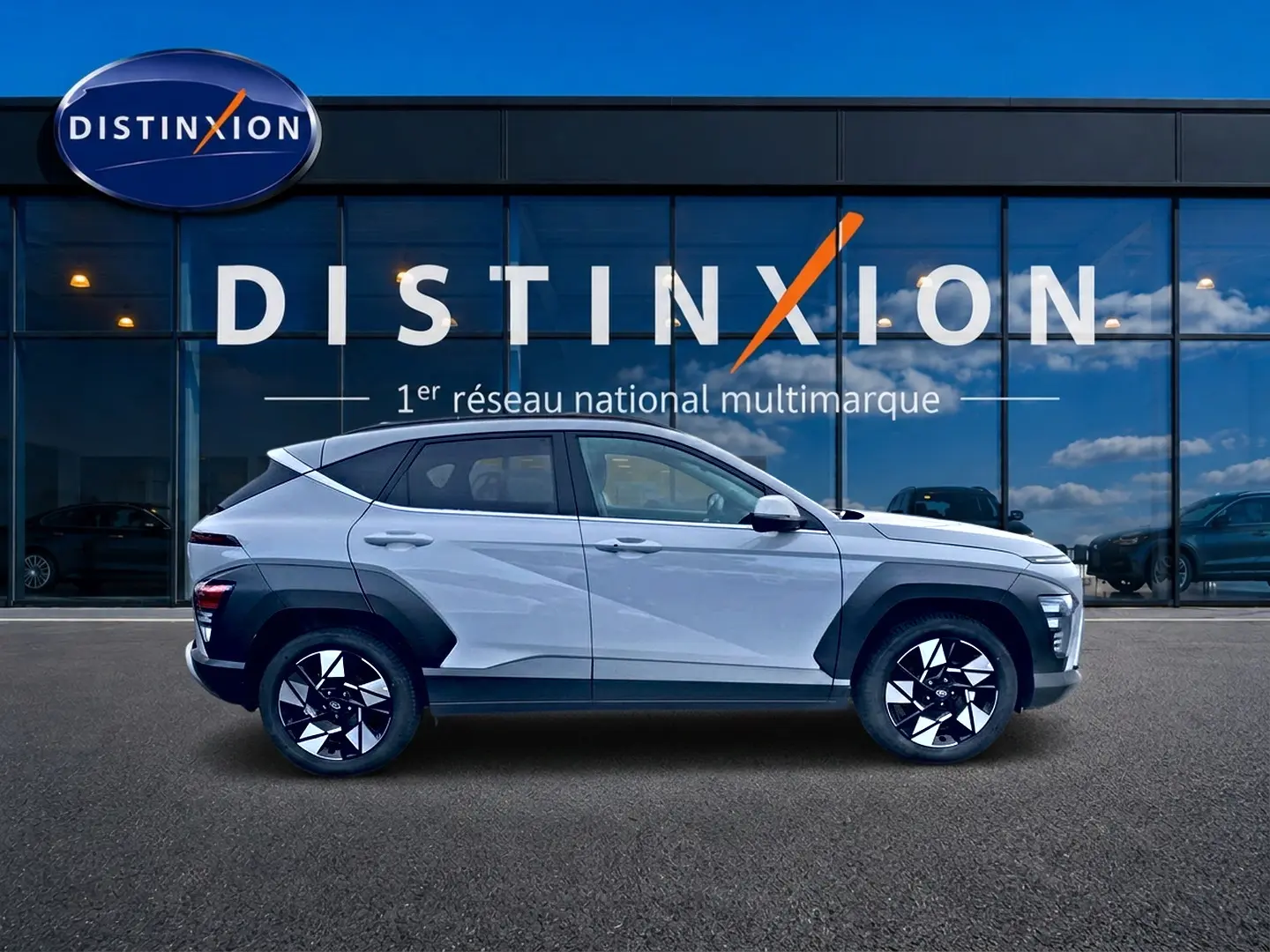 Profil côté gauche d'un Hyundai Kona gris 2025 avec protections noires et jantes alliage bicolores devant un showroom Distinxion.