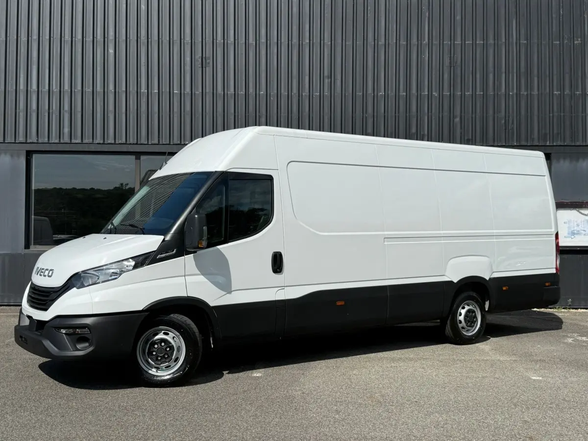 Fourgon Iveco Daily blanc vu de profil côté gauche, avec pare-chocs et bas de caisse noirs, stationné devant un bâtiment industriel.