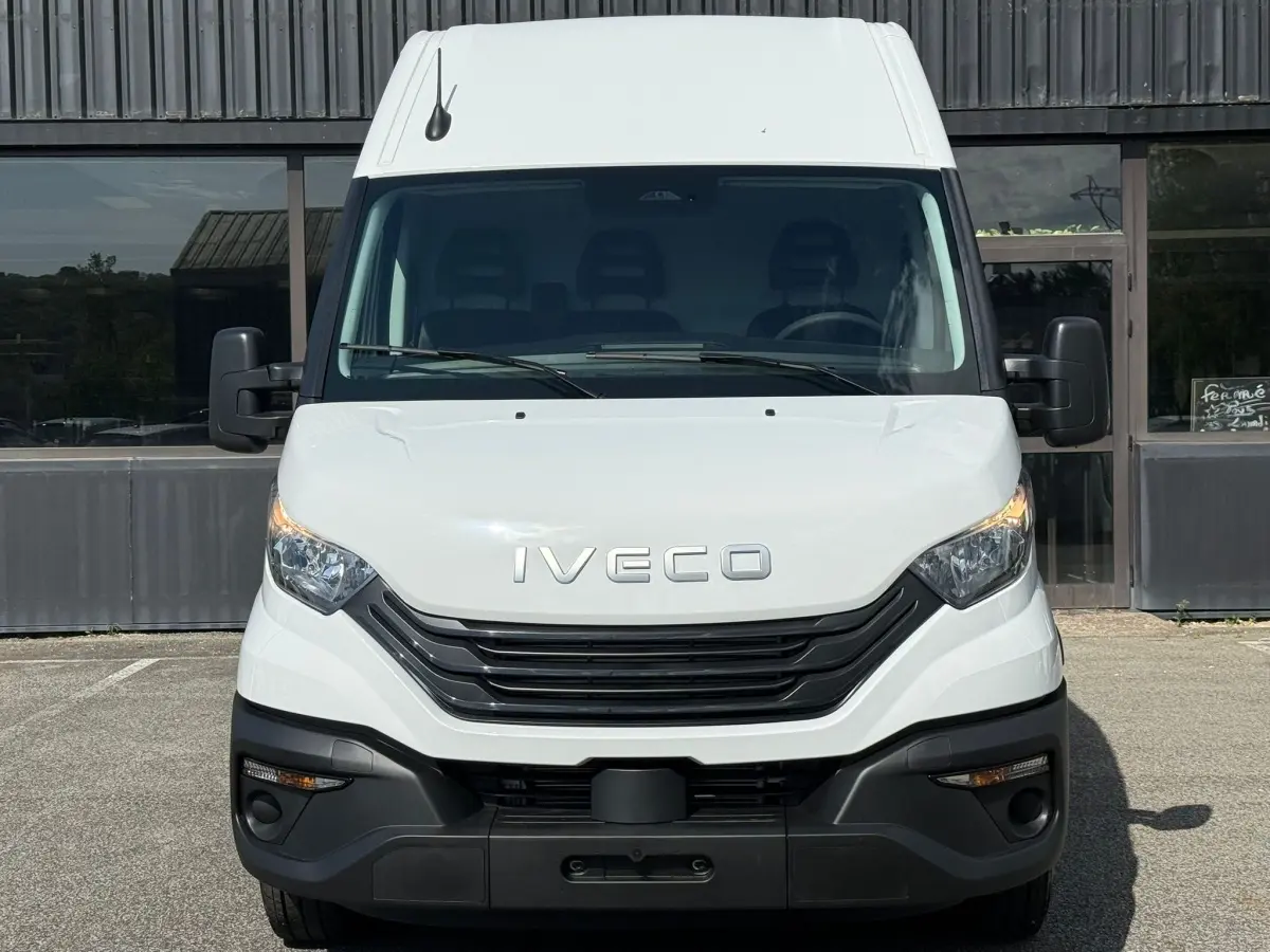 Vue frontale d’un utilitaire Iveco Daily blanc avec calandre noire et phares allumés, stationné devant un bâtiment.