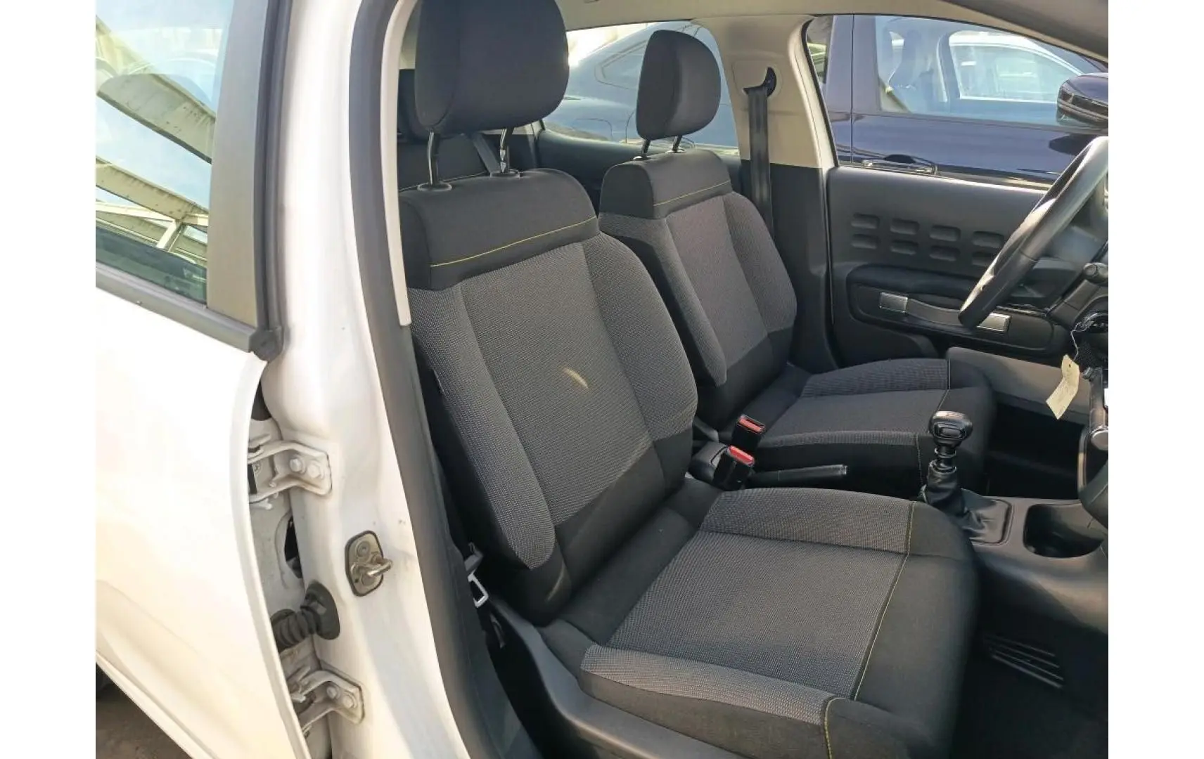 Vue intérieure côté conducteur d'une Citroën C3 blanche 2021, montrant les sièges avant en tissu gris avec surpiqûres jaunes.