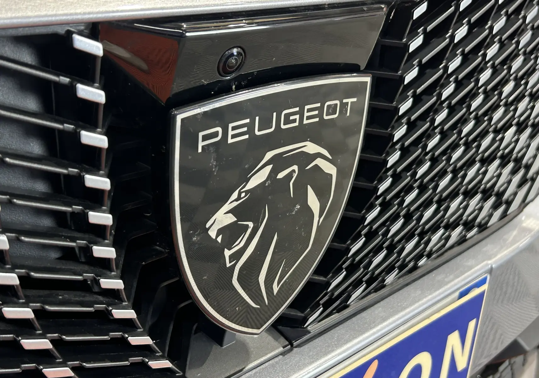 Gros plan sur le logo Peugeot noir et argent sur la calandre gris Artense du Peugeot 3008 III Hybrid 145 e-DSC6 Allure.
