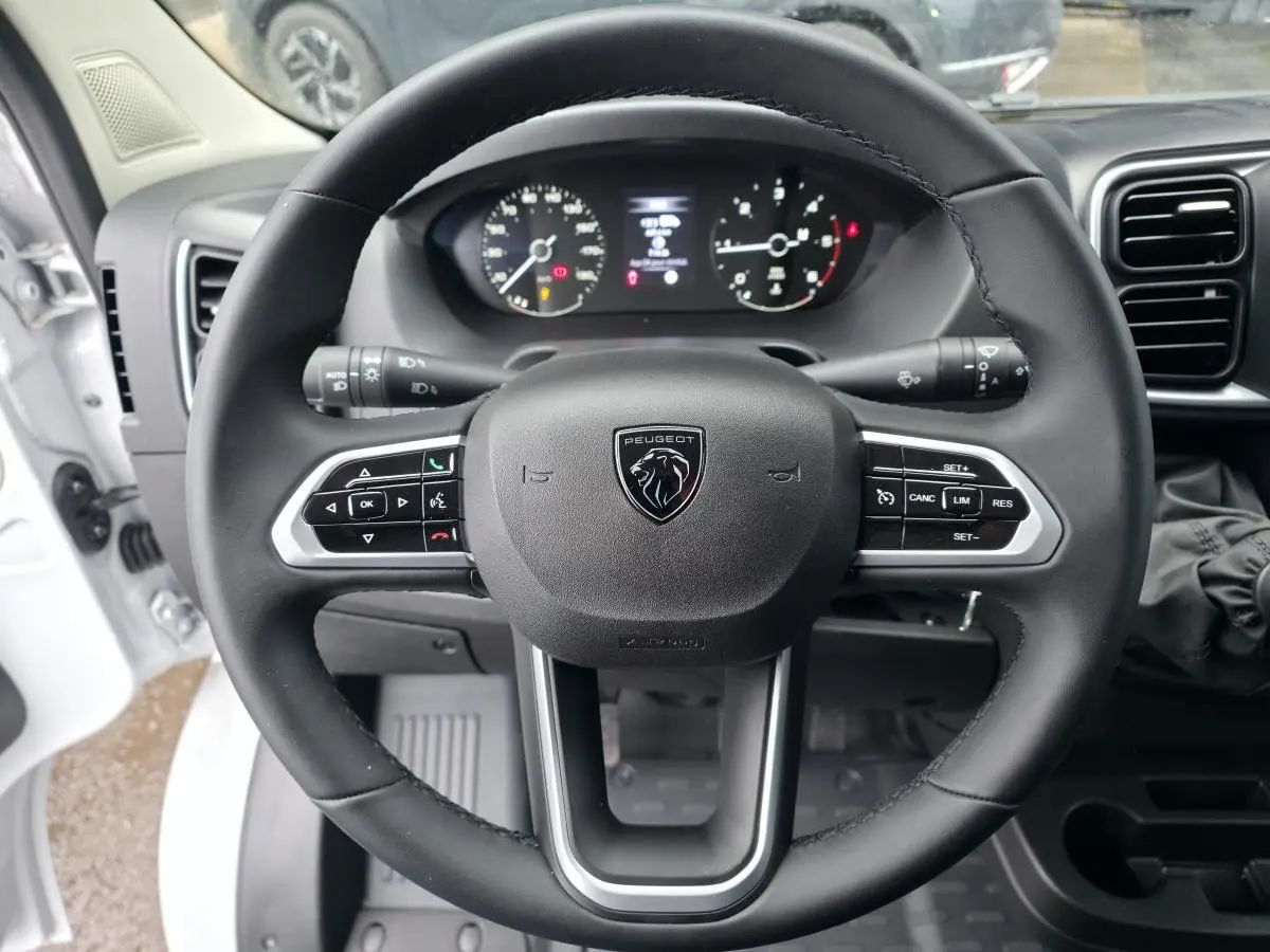 Vue rapprochée du volant cuir noir avec commandes intégrées du Peugeot Boxer Fourgon blanc, tableau de bord et compteurs en arrière-plan.