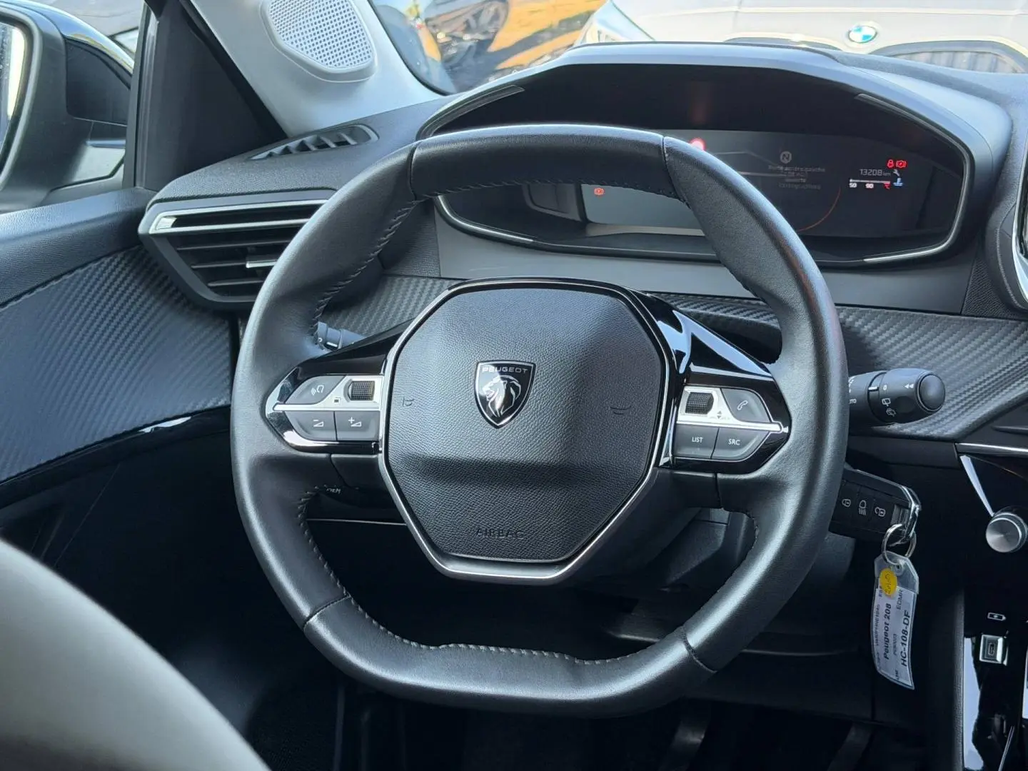 Vue rapprochée du volant compact noir avec logo Peugeot au centre et tableau de bord numérique en arrière-plan.